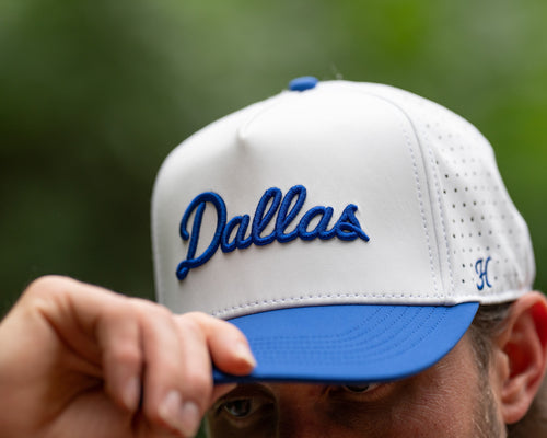 White and blue cap with 'Dallas' text held by a person against a blurred green background