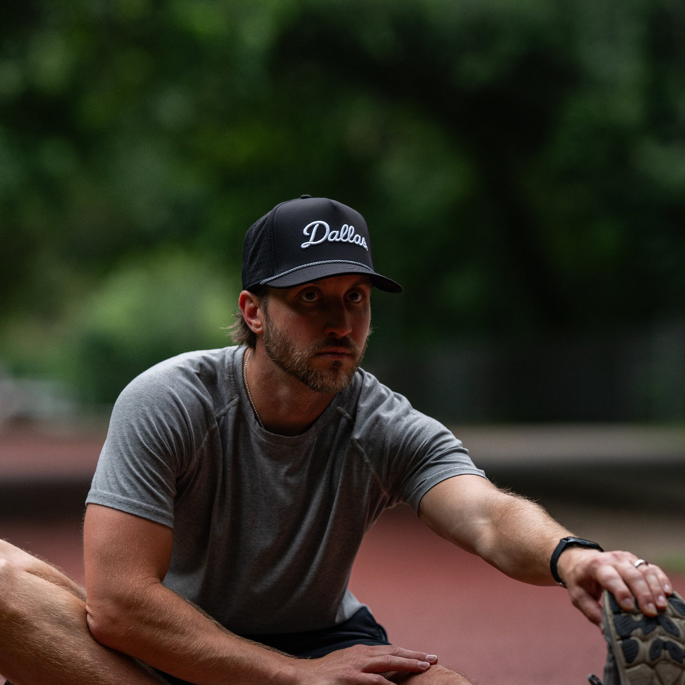 Man wearing a cap with 'Dallas' text, sitting outdoors with blurred greenery in the background