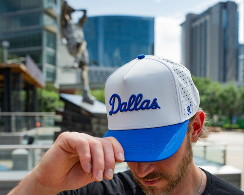 Man wearing a white and blue cap with 'Dallas' printed on it, standing in an urban setting.
