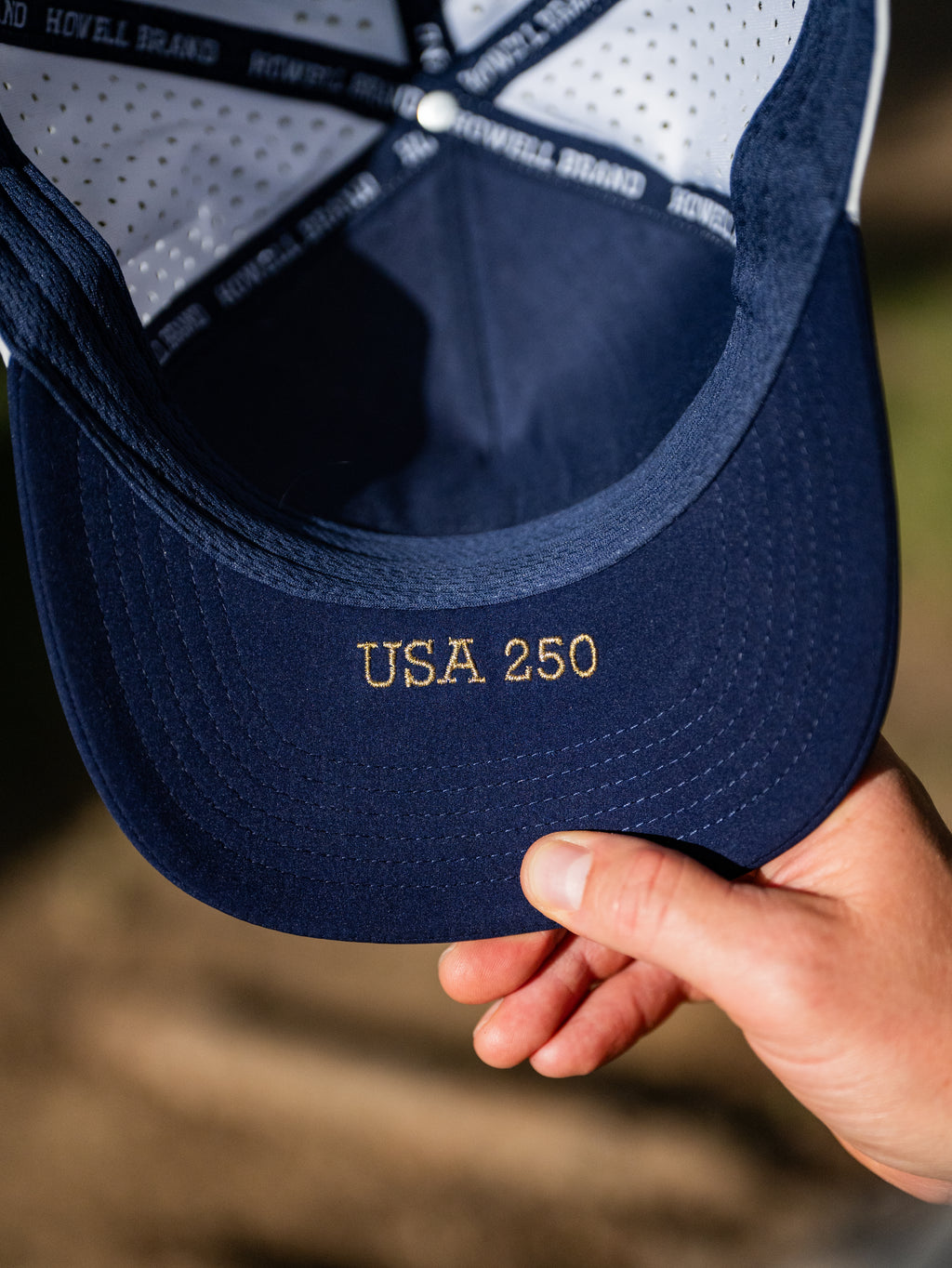 Navy blue cap with 'USA 250' embroidery held by a hand against a blurred background