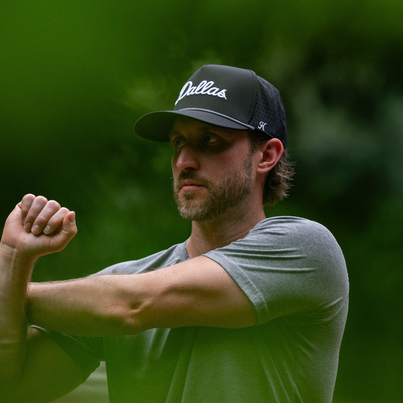 Man wearing a black cap with 'Dallas' branding, standing outdoors with greenery in the background.